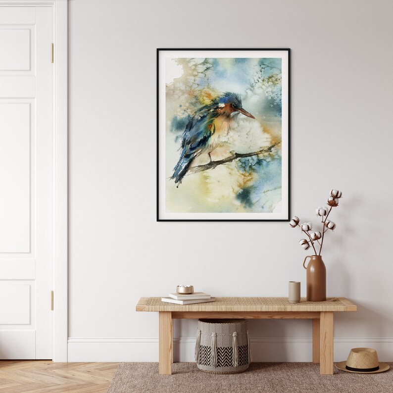 May include: Watercolor painting of a bird perched on a branch, framed in black. The bird features blue, yellow, and brown hues. The background has soft, blended colors of blue, yellow, and beige. The artwork is displayed on a white wall.