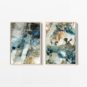 May include: Two framed watercolor paintings. The artwork features abstract botanical designs in shades of blue, brown, and beige. The paintings have a light wood frame and are displayed against a white background.