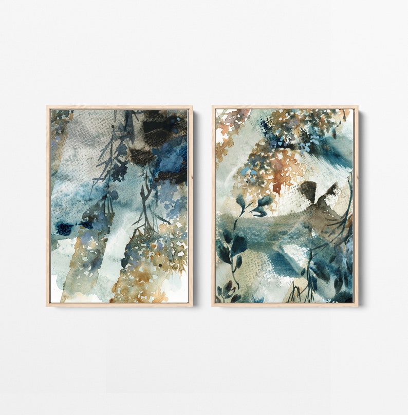 May include: Two framed watercolor paintings. The artwork features abstract botanical designs in shades of blue, brown, and beige. The paintings have a light wood frame and are displayed against a white background.
