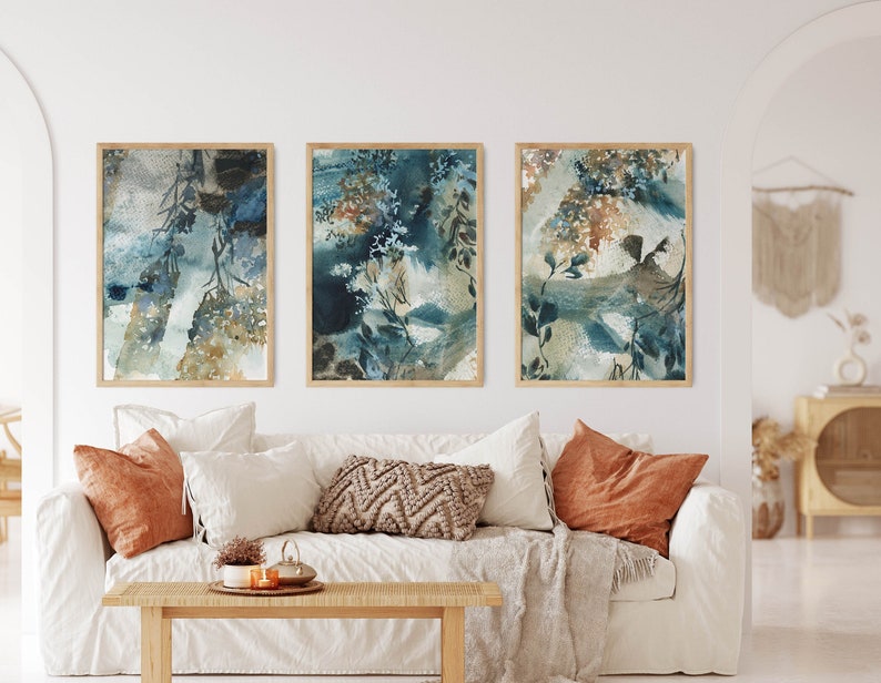 K&ouml;nnte beinhalten: Drei gerahmte abstrakte Aquarelldrucke. Das Kunstwerk zeigt eine Mischung aus Blau-, T&uuml;rkis- und Braunt&ouml;nen mit botanischen und strukturierten Elementen. Die Drucke werden &uuml;ber einem wei&szlig;en Sofa mit orangefarbenen und neutralfarbenen Kissen pr&auml;sentiert.