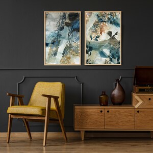 May include: Two framed watercolor art prints with abstract botanical designs in shades of blue, brown, and white. The prints are displayed on a dark gray wall above a mid-century modern wooden cabinet and a yellow armchair.