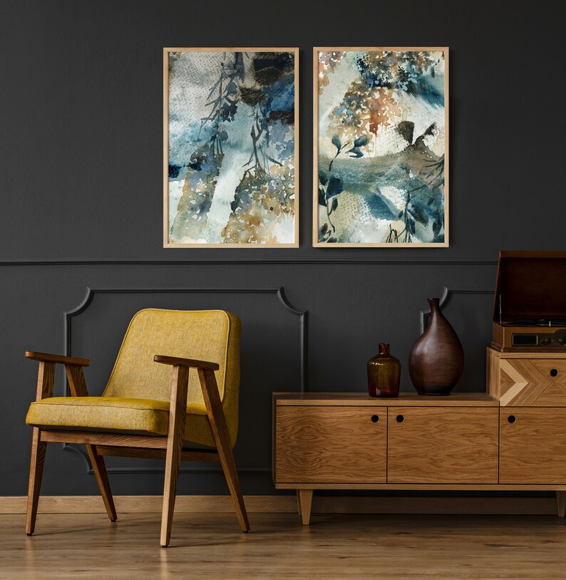 May include: Two framed watercolor art prints with abstract botanical designs in shades of blue, brown, and white. The prints are displayed on a dark gray wall above a mid-century modern wooden cabinet and a yellow armchair.