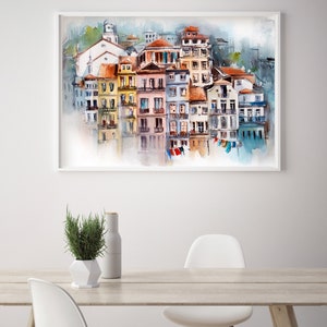 May include: Watercolor painting of a European coastal town with colorful buildings, red tile roofs, and a church. The artwork is framed in white and hangs on a white wall. The painting is reflected in the water below. A table with a plant and chairs is in the foreground.