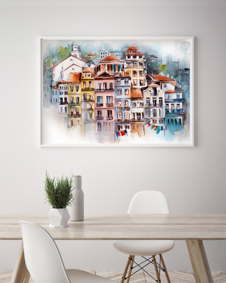 May include: Watercolor painting of a European coastal town with colorful buildings, red tile roofs, and a church. The artwork is framed in white and hangs on a white wall. The painting is reflected in the water below. A table with a plant and chairs is in the foreground.