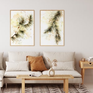 May include: Two framed watercolor prints of pine branches. The artwork features green and brown pine needles against a soft, abstract background with yellow and beige tones. The frames are a light wood color.