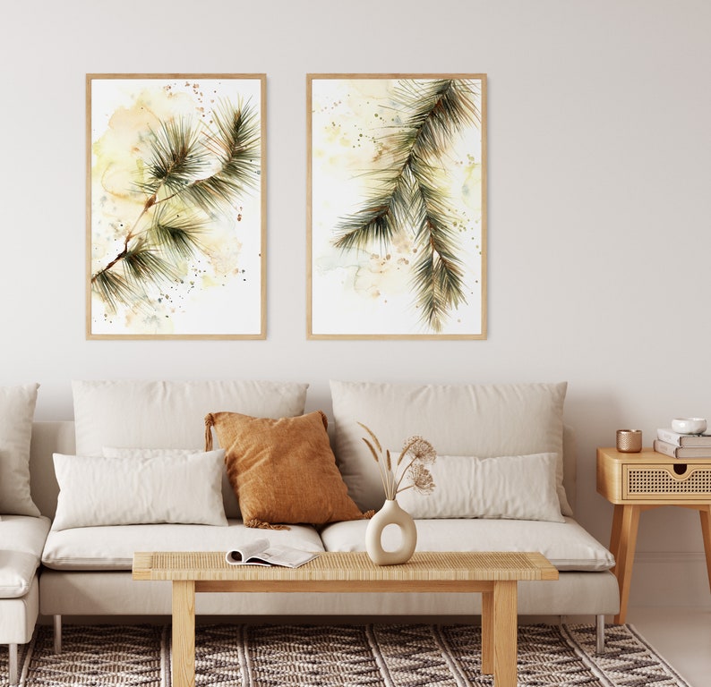 May include: Two framed watercolor prints of pine branches. The artwork features green and brown pine needles against a soft, abstract background with yellow and beige tones. The frames are a light wood color.