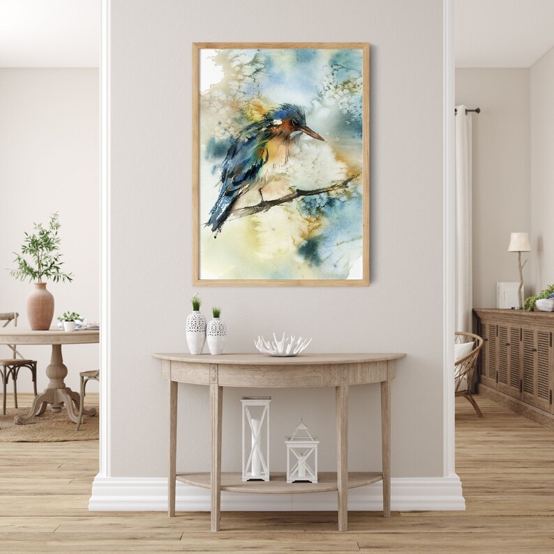 May include: Watercolor painting of a bird perched on a branch, framed in light wood. The bird features blue, green, and brown hues, set against a soft, abstract background of blues and yellows. The artwork is displayed in a well-lit interior setting.