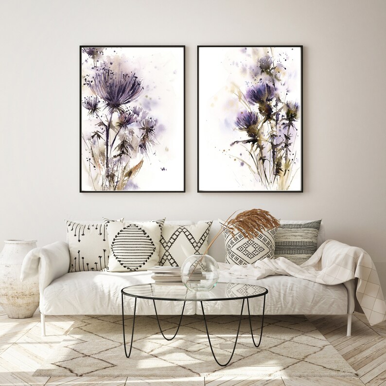 May include: Two framed watercolor paintings of purple thistle flowers. The artwork features a white background with splashes of color and black frames. The paintings are displayed above a white sofa with decorative pillows.