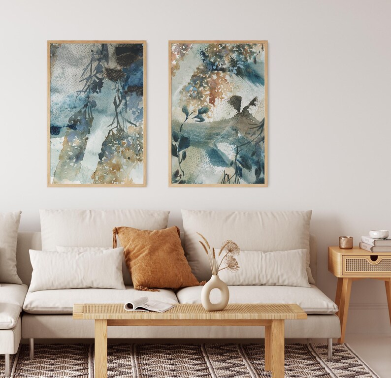 May include: Two framed watercolor paintings with botanical themes. The artwork features blue, brown, and beige tones, with a focus on floral and leaf motifs. The paintings are displayed above a cream-colored sofa with decorative pillows, a wooden coffee table, and a woven rug.