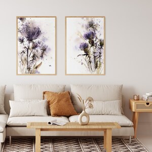 May include: Two framed watercolor prints of purple thistle flowers. The artwork features delicate brushstrokes and a soft color palette of purple, brown, and white. The prints are displayed above a beige sofa in a living room setting.