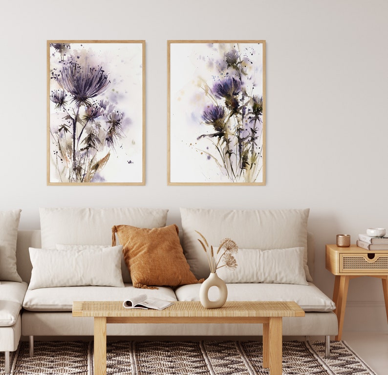 May include: Two framed watercolor prints of purple thistle flowers. The artwork features delicate brushstrokes and a soft color palette of purple, brown, and white. The prints are displayed above a beige sofa in a living room setting.