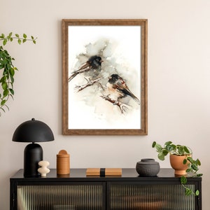 May include: A framed watercolour print depicting two birds on branches. The artwork uses a neutral colour palette with brown, black, and white tones. The frame is wooden. The print is displayed on a white wall above a black cabinet.