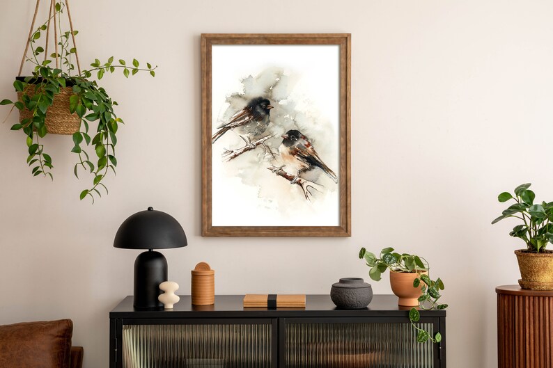 May include: A framed watercolour print depicting two birds on branches. The artwork uses a neutral colour palette with brown, black, and white tones. The frame is wooden. The print is displayed on a white wall above a black cabinet.