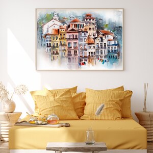 May include: Watercolor painting of a European coastal town with colorful buildings and red-tiled roofs. The artwork is framed and hangs above a yellow daybed with pillows. A small wooden table and decorative items complete the scene.