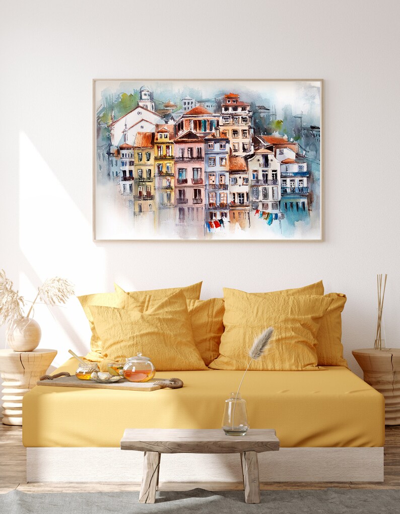 May include: Watercolor painting of a European coastal town with colorful buildings and red-tiled roofs. The artwork is framed and hangs above a yellow daybed with pillows. A small wooden table and decorative items complete the scene.