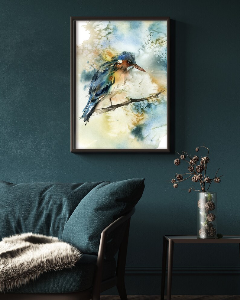 May include: Watercolor painting of a bird perched on a branch, framed in a black frame. The bird features blue, orange, and brown hues. The background has soft watercolor washes of blue, yellow, and white. The artwork is displayed on a dark teal wall.
