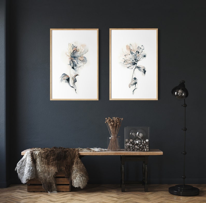 Puede incluir: Dos impresiones enmarcadas de acuarela floral con marcos de madera clara cuelgan de una pared azul oscuro. La obra de arte presenta flores abstractas en tonos de azul, beige y gris. Debajo de las obras de arte hay un banco de madera con una manta de piel y objetos decorativos.