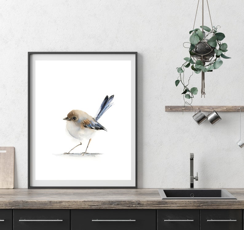 Puede incluir: Una impresi&oacute;n en acuarela enmarcada de un p&aacute;jaro peque&ntilde;o con cabeza marr&oacute;n, cuerpo blanco y plumas de cola azules. El p&aacute;jaro est&aacute; sobre un fondo blanco. El marco es negro. La impresi&oacute;n se exhibe sobre una encimera de madera.