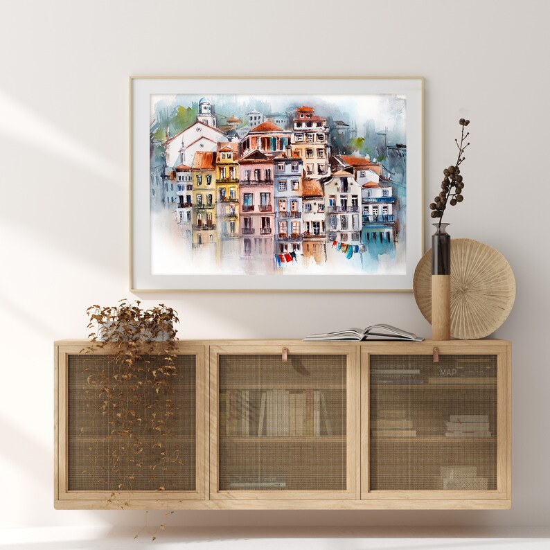 May include: A framed watercolor print of a coastal town with colorful buildings and a church. The artwork is displayed above a wooden cabinet with woven doors, complemented by decorative items like a vase and a round wall hanging.