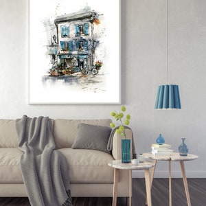 May include: Framed watercolour print depicting a European building with blue shutters, a flower shop, and a bicycle. The artwork hangs above a beige sofa with a grey throw and cushions. Two small side tables with decor are in front of the sofa.