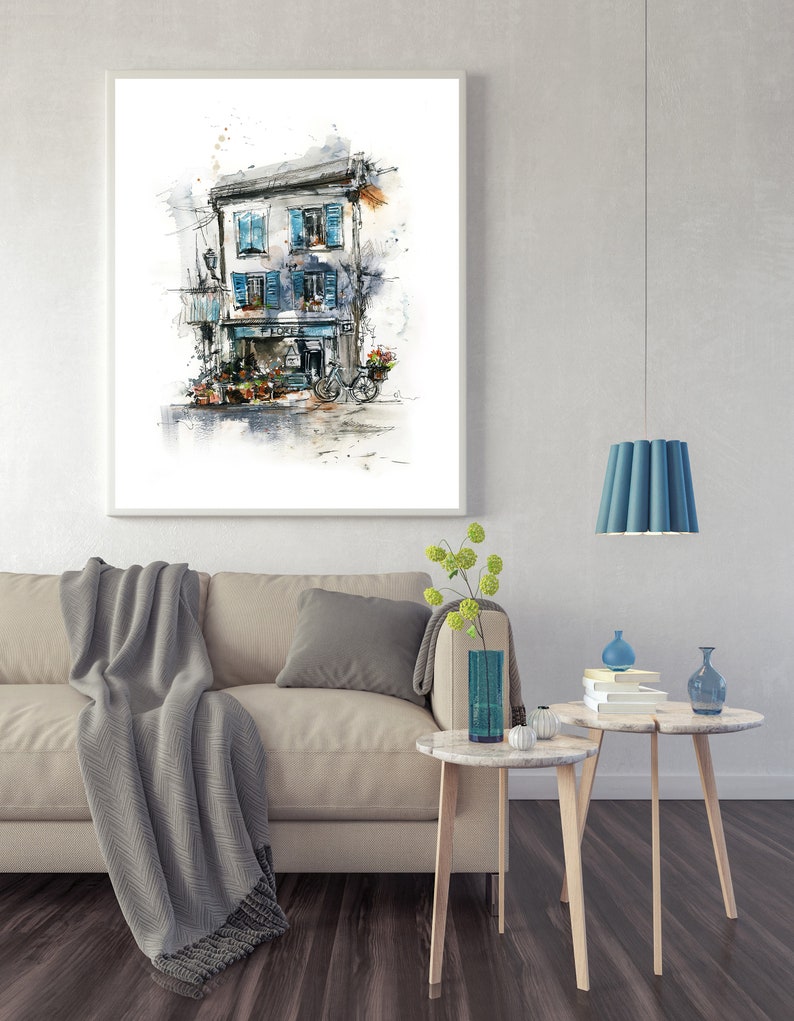 May include: Framed watercolour print depicting a European building with blue shutters, a flower shop, and a bicycle. The artwork hangs above a beige sofa with a grey throw and cushions. Two small side tables with decor are in front of the sofa.