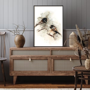May include: A framed watercolour print depicting two birds on branches. The birds have black and brown feathers, set against a soft, neutral backdrop. The artwork is displayed on a wooden cabinet with a woven front, adding a touch of rustic charm to the room.