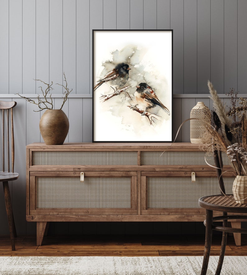 May include: A framed watercolour print depicting two birds on branches. The birds have black and brown feathers, set against a soft, neutral backdrop. The artwork is displayed on a wooden cabinet with a woven front, adding a touch of rustic charm to the room.