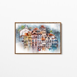 May include: Watercolor painting of a European cityscape with colorful buildings and red-tiled roofs. The artwork is framed in a wooden frame and depicts a waterfront scene with reflections in the water. The buildings are painted in shades of yellow, pink, and blue.