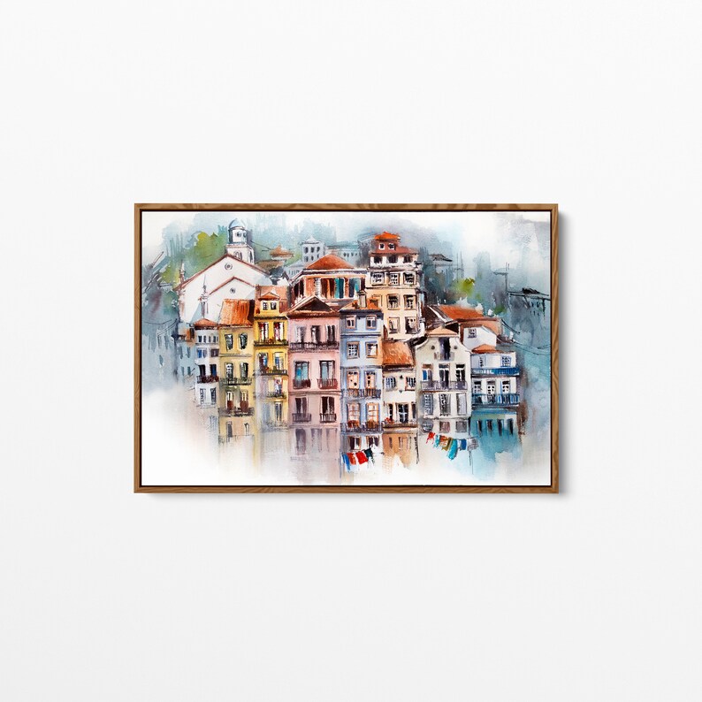 May include: Watercolor painting of a European cityscape with colorful buildings and red-tiled roofs. The artwork is framed in a wooden frame and depicts a waterfront scene with reflections in the water. The buildings are painted in shades of yellow, pink, and blue.