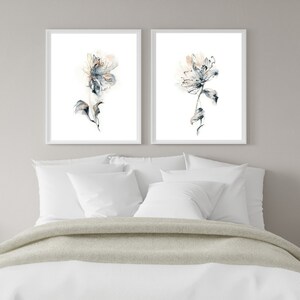 Puede incluir: Dos impresiones florales en acuarela enmarcadas. Cada impresi&oacute;n presenta una flor delicada y abstracta en tonos azules, melocot&oacute;n y grises sobre un fondo blanco. Las impresiones se exhiben sobre una cama con ropa de cama blanca y una manta gris claro.