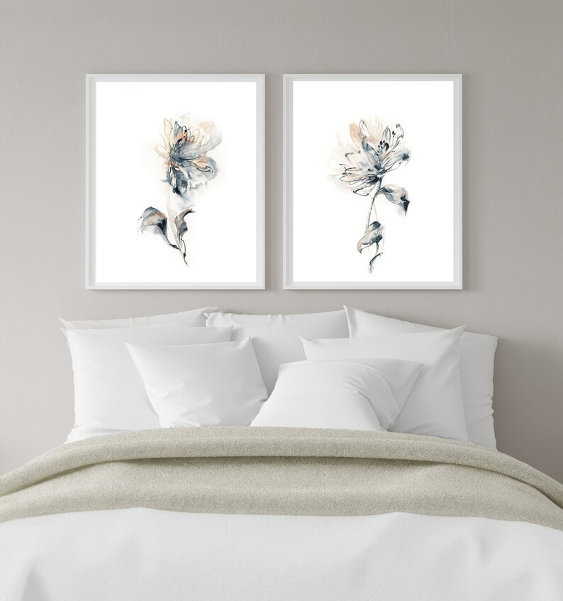 Puede incluir: Dos impresiones florales en acuarela enmarcadas. Cada impresi&oacute;n presenta una flor delicada y abstracta en tonos azules, melocot&oacute;n y grises sobre un fondo blanco. Las impresiones se exhiben sobre una cama con ropa de cama blanca y una manta gris claro.