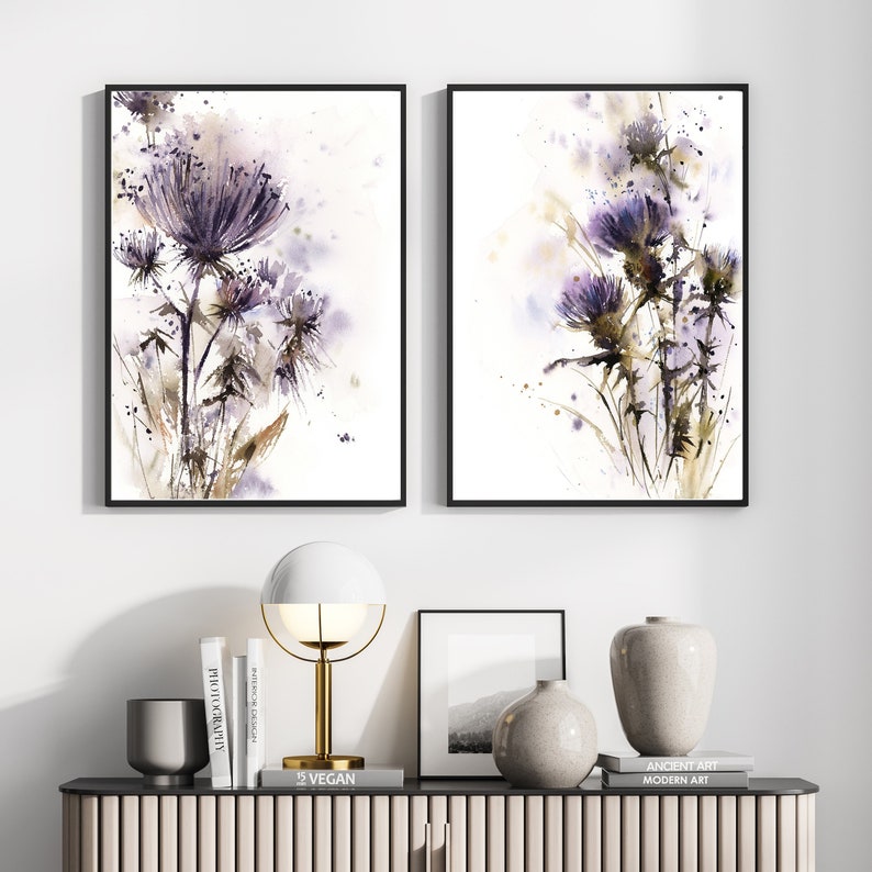 May include: Two framed watercolor prints featuring purple thistle flowers. The artwork has a soft, painterly style with splashes of color against a white background. The frames are black, and the overall aesthetic is modern and botanical.