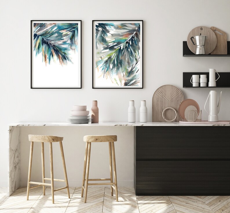 Puede incluir: Dos pinturas de acuarela enmarcadas de follaje abstracto azul y verde cuelgan sobre una encimera de cocina. Las pinturas tienen marcos negros y se exhiben en una pared blanca. Dos taburetes de madera se encuentran frente a la encimera, y hay objetos decorativos.