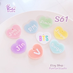 Puede incluir: Un conjunto de siete dijes en forma de corazón de color pastel con letras blancas. Los dijes presentan los nombres de los miembros de la banda de chicos surcoreana BTS: RM, Jin, Suga, J-Hope, Jimin, V y Jungkook. Los dijes están dispuestos sobre una superficie blanca con el texto "S61" y "Etsy Shop: Punfun Studio" visible.