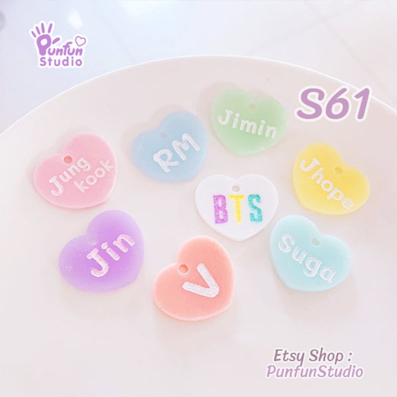 May include: A collection of eight heart-shaped charms in pastel colors, each featuring white text including names like Jungkook, RM, Jimin, Jhope, Jin, V, Suga, and the letters BTS in multicolored font. Each charm has a small hole at the top.