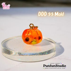 May include: A small orange pumpkin-shaped resin charm with a cute face, hanging from a gold chain. The charm is on a clear, glittery surface. DDD 33 Mold.