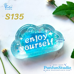 Puede incluir: Un adorno en forma de nube de resina azul con purpurina y las palabras "enjoy yourself" en su interior. El adorno está sobre un fondo blanco.