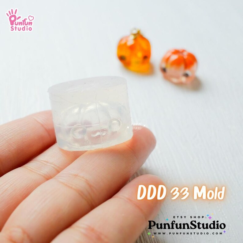 May include: A clear silicone mold for making miniature pumpkin charms. The mold is in the shape of a pumpkin and has a detailed design. The mold is being held in a hand. The text "DDD 33 Mold" is visible in the image.