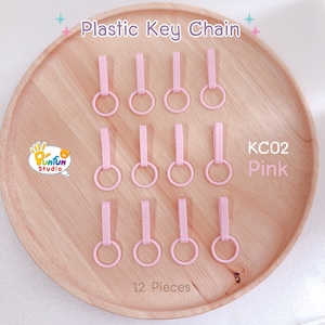 May include: Twelve pink plastic keychains with a metal ring. The keychains are arranged in three rows of four on a wooden surface. The text "KC02 Pink" is visible in the image.