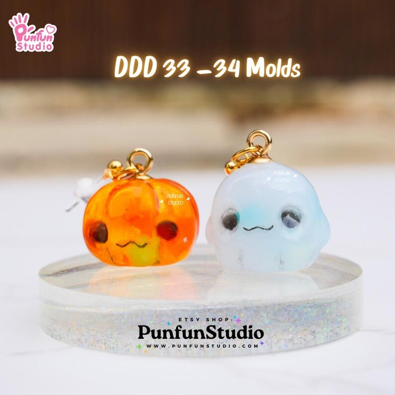 May include: Two Halloween-themed resin charms, a pumpkin and a ghost, with gold accents and cute faces. The charms are on a clear display stand. DDD 33-34 Molds.