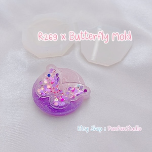 May include: A purple butterfly resin mold with glitter embedded in the resin. The mold is on a white surface with two clear silicone molds in the background. The text "R269 x Butterfly Mold" is written in pink on the silicone molds.