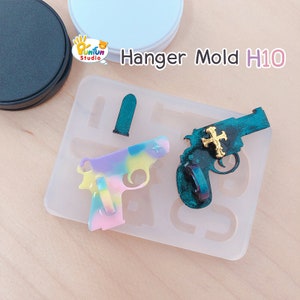 May include: A clear silicone mold with two gun-shaped cavities. One gun is filled with a multi-colored resin, and the other is filled with a dark green resin with a gold cross. The mold is labeled "Hanger Mold H10".