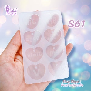 May include: A translucent, rectangular silicone mold featuring eight heart-shaped cavities. Each heart contains a small hole at the top and embossed text including Jin, BTS, Suga, Jung Kook, Jimin, RM, V, and J-Hope. The mold is flexible and clear.
