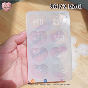 May include: A rectangular, translucent silicone mold featuring eight heart-shaped cavities, each embossed with the text BTS. The mold is labeled S61/2 Mold at the top. Several finished, white, heart-shaped pieces with the text BTS are visible nearby.