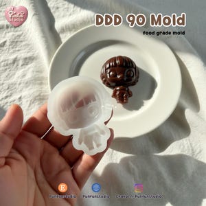 K&ouml;nnte beinhalten: Eine durchscheinende Silikonform in Form einer stilisierten Figur mit Bob-Frisur, begleitet von einer dunkelbraunen Schokoladenfigur, die aus derselben Form gegossen wurde. Der Text DDD 90 Mold und food grade mold ist oben im Bild zu sehen.