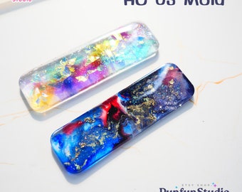 Pode incluir: Dois moldes retangulares de resina com um design inspirado na galáxia. O molde à esquerda tem um fundo azul claro, rosa e amarelo com flocos dourados. O molde à direita tem um fundo azul escuro com detalhes vermelhos, brancos e dourados. O texto "HC 03 Mold" está impresso em preto no topo da imagem.
