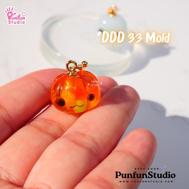 May include: A small orange pumpkin-shaped resin charm with a gold loop for hanging. The charm has a cute cartoon face with black eyes and a yellow mouth. The text "DDD 33 Mold" is visible in the background.
