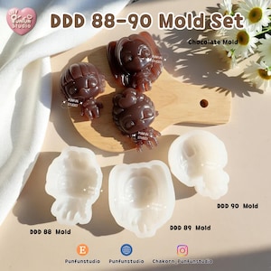K&ouml;nnte beinhalten: Ein DDD 88-90 Formen-Set mit drei braunen Schokoladenfiguren in Form stilisierter Charaktere und drei dazugeh&ouml;rigen wei&szlig;en Silikonformen mit der Beschriftung DDD 88 Mold, DDD 89 Mold und DDD 90 Mold auf einer hellen Oberfl&auml;che.