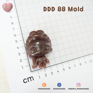 K&ouml;nnte beinhalten: Eine braune, durchscheinende, stilisierte Puppenfigur-Form mit der Aufschrift DDD 88 Mold. Die Figur hat einen gro&szlig;en Kopf mit detailliertem Haar und Gesichtsz&uuml;gen und liegt auf einer Rastermatte mit Zentimeterangaben, die eine H&ouml;he von 4 cm zeigt.