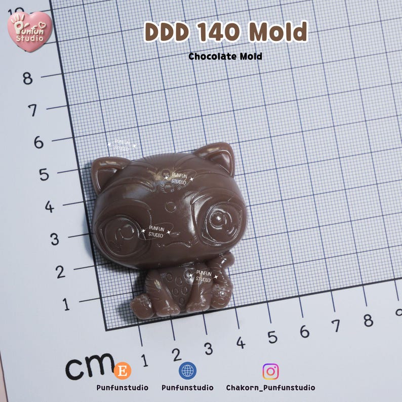 K&ouml;nnte beinhalten: Eine braune Schokoladenform in Form einer stilisierten Katze mit gro&szlig;en Augen, platziert auf einer Rastermatte mit Zentimeterangaben. Der Text DDD 140 Mold und Chocolate Mold ist oben aufgedruckt. Die Form ist etwa 3,5 cm breit.